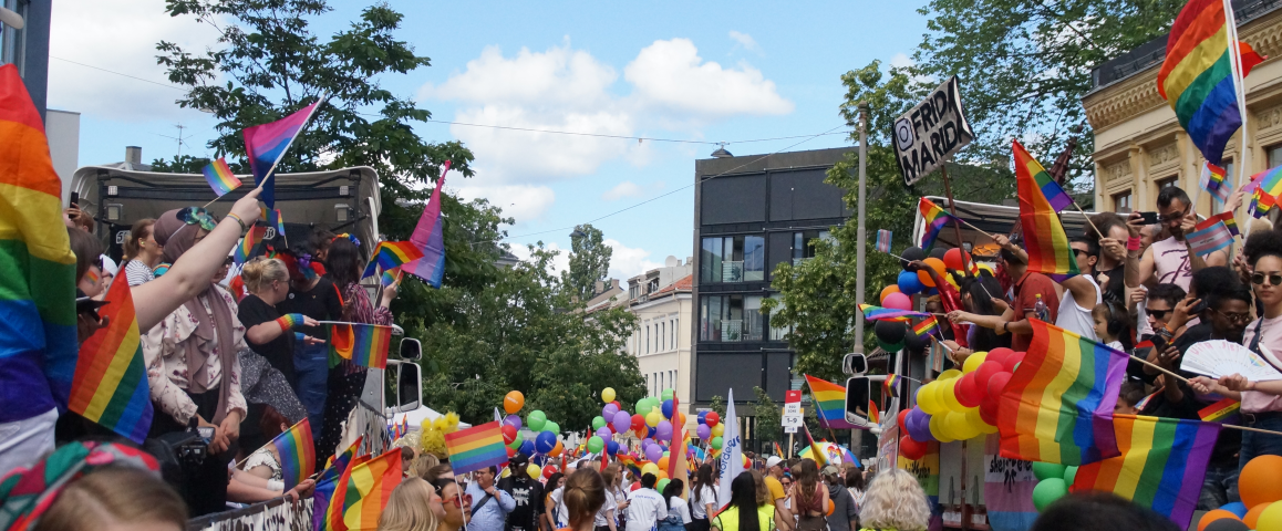 Bilde fra forrige parade. "Vår trailer" på den ene siden vinker til forbipasserende trailer, og i forgrunnen sees masse mennesker og prideflagg og ballonger.