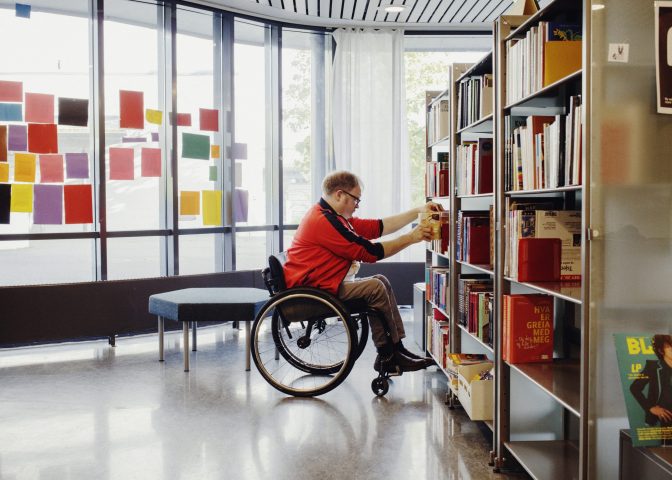 Mann i rullestol setter bøker inn i bibliotekhylle