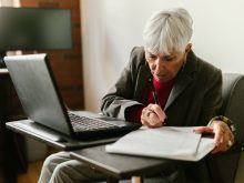 an elderly woman writing on paper near the laptop