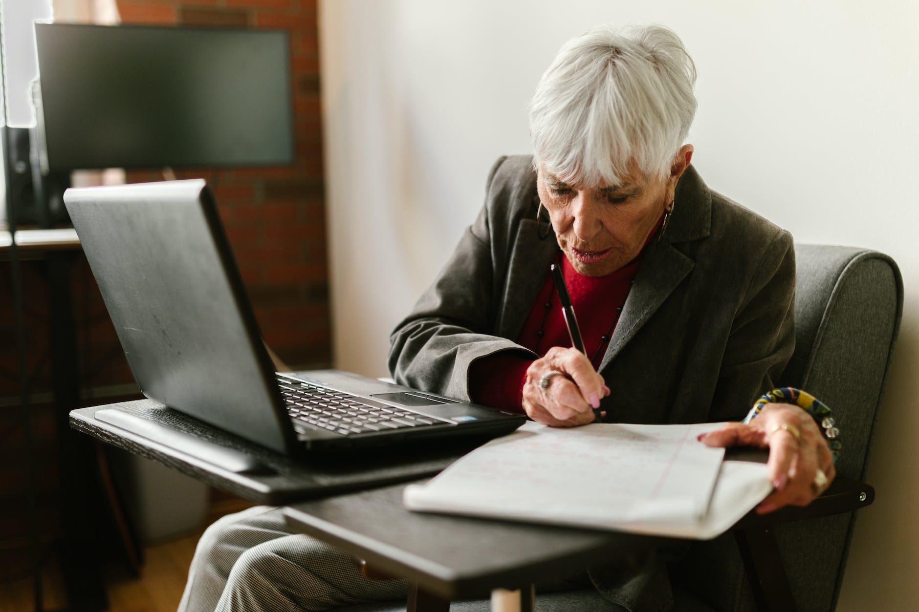 an elderly woman writing on paper near the laptop