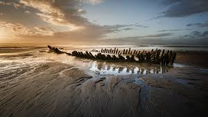Berrow Beach A wrecked boat sits on a sandy beach at sunset, with the sky filled with orange and grey clouds.