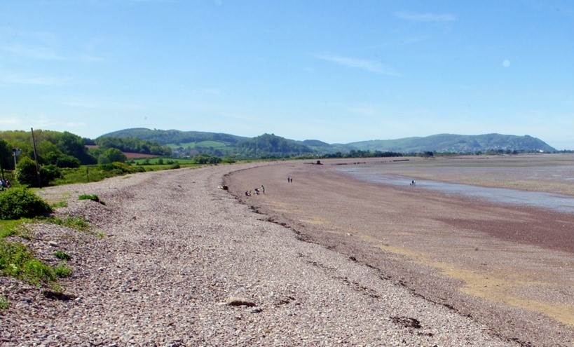Blue Anchor Bay A pebble beach extends into a shallow bay under a blue sky, with a green hillside visible in the distance. | Blue Anchor Bay