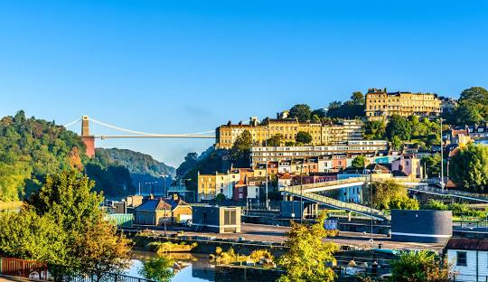 Bristol Clifton Suspension Bridge spans the Avon Gorge in Bristol, with colourful buildings lining the hills under a clear blue sky.