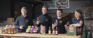 3 men and a lady stand behind a wooden bar, all holding glasses full of cider. On the bar is a display of Harry's Cider produce.