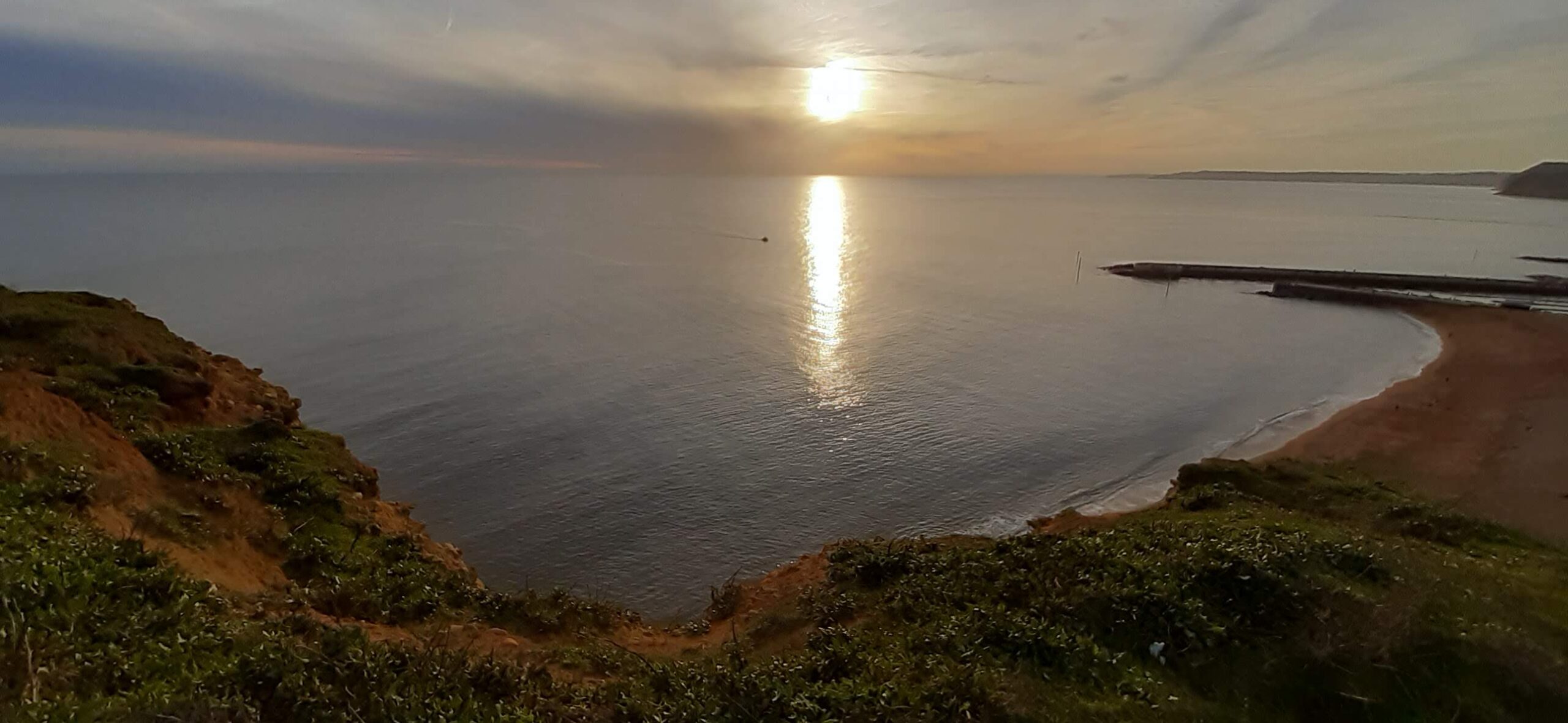 West Bay A coastal view shows a cliff overlooking the sea at sunset, with the sky filled with orange and grey clouds. | Jurassic Coastline