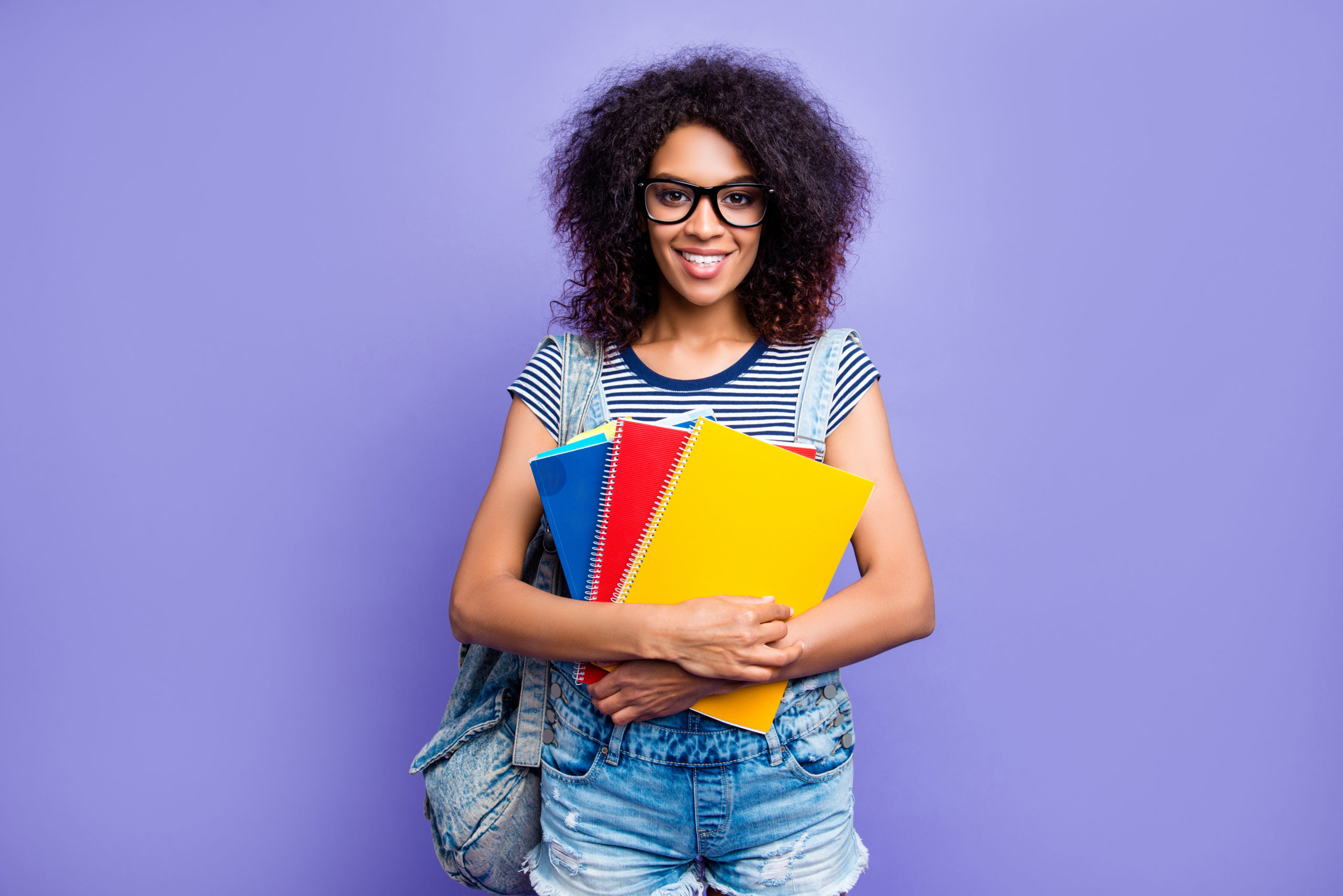 Portrait of smart educated girl with beaming smile in denim outfit eyewear having colorful textbooks in hands looking at camera isolated on violent background. Training skills high-school concept