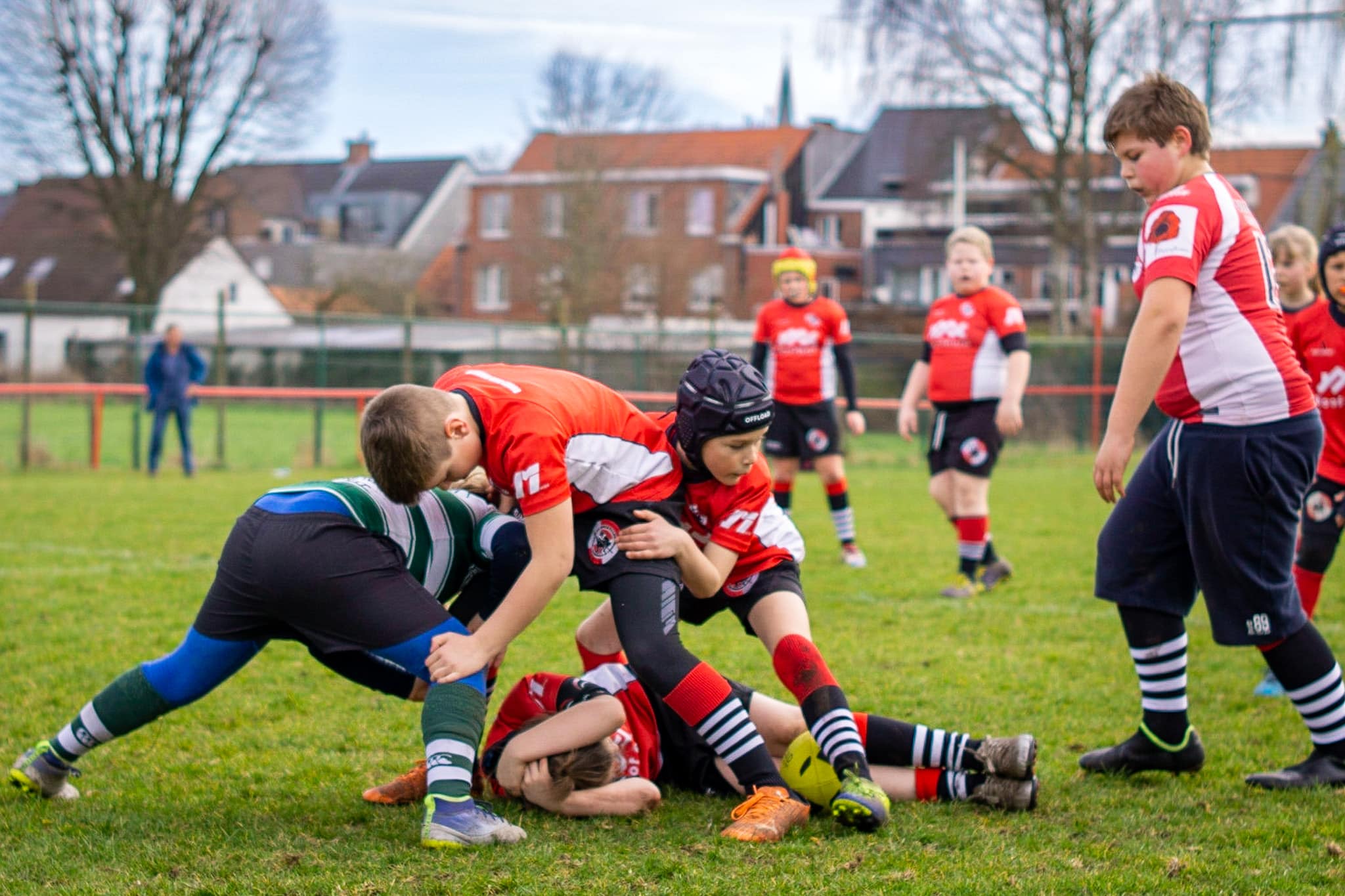 Kinderen spelen een intensieve rugbywedstrijd, waarbij een speler in het groen-wit wordt getackeld door twee spelers van het rode team. Een gele rugbybal ligt op de grond.