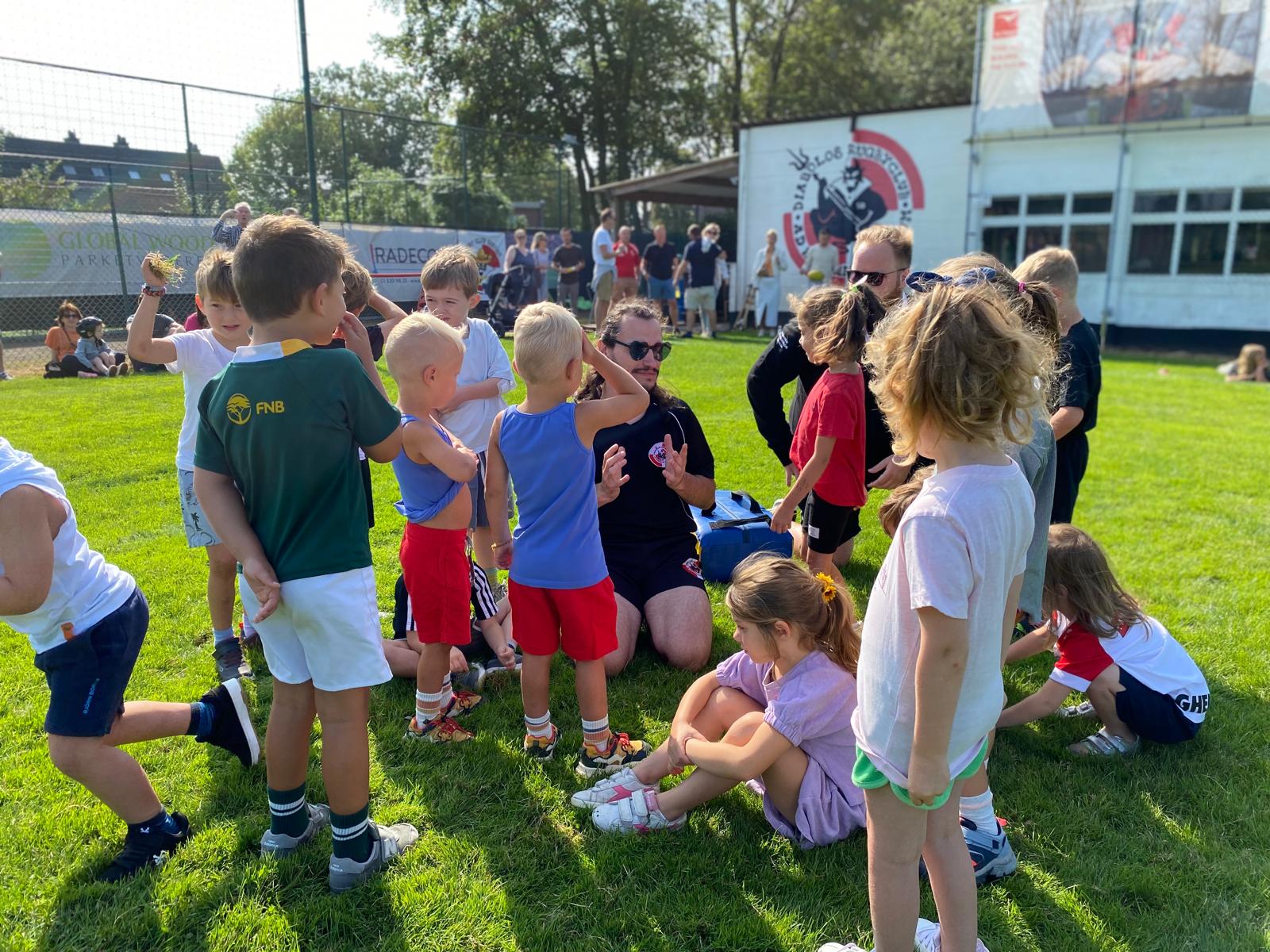 Een groep jonge kinderen speelt en luistert naar een coach op het veld van Rugby Club Diabolos Schilde tijdens een zonnige dag.