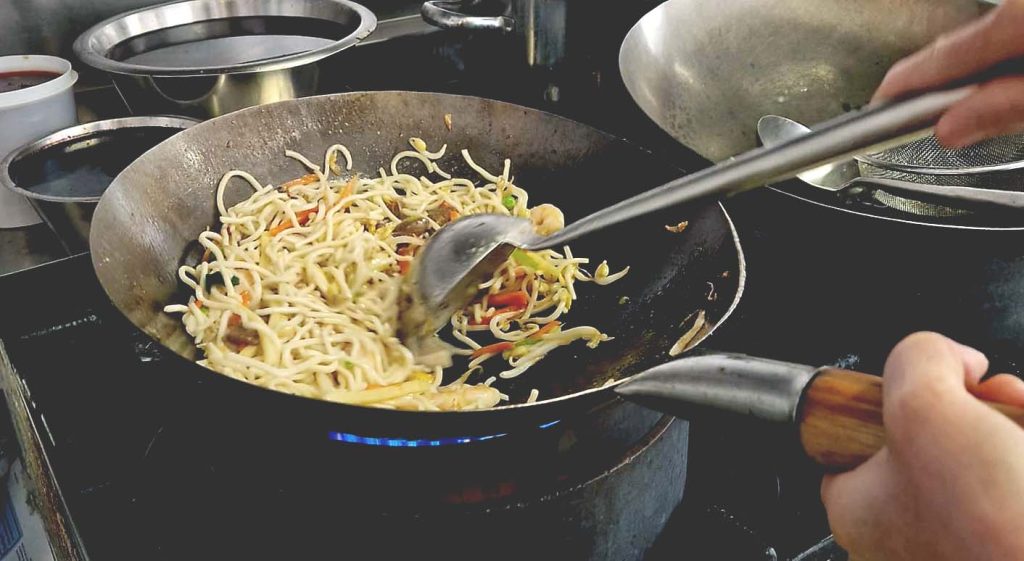 A close-up photo of a wok pan with fried noodles and vegetables being stirred with a ladle.