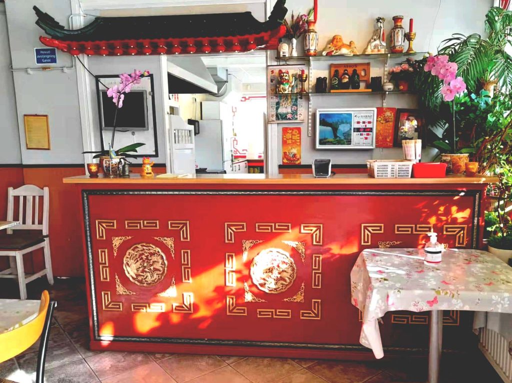 Photo of Restaurant Ming's counter that is red with golden patterns in Chinese style, green plants and flowers in shop windows next to and on the counter. Chinese decor.