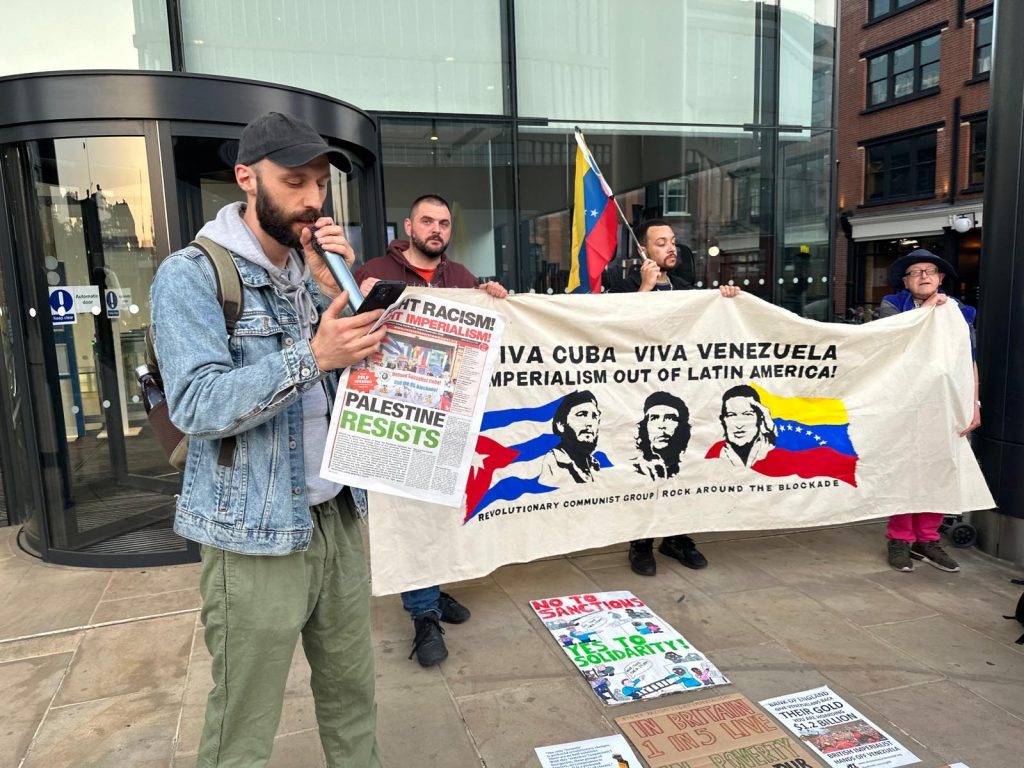 A man speaks into a microphone, in his other hand he holds a copy of the newspaper Fight Racism! Fight Imperialism! In the background three people hold a banner reading 'Viva Cuba, Viva Venezuela, Imperialism out of Latin America' above images of Fidel Castro, Che Guevara and Hugo Chavez.