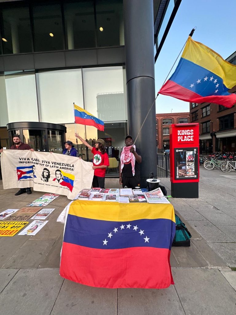 A man waves a Venezuela flag, standing behind a stall tabel in front of the Guardian offices, London