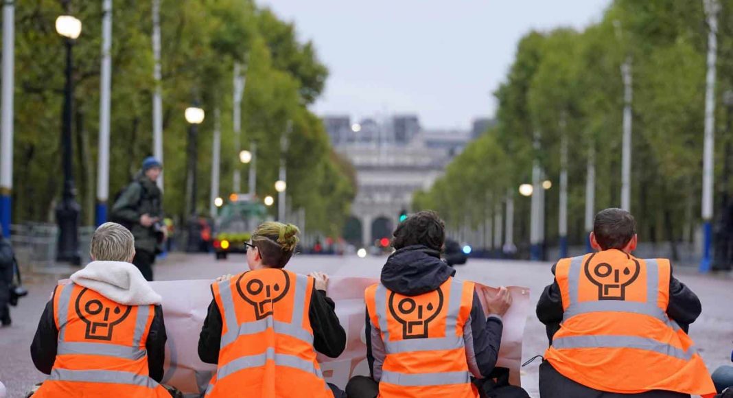 Four people in orange hi-viz vests with the Just Stop Oil logo sit with their back facing the camera on a road.