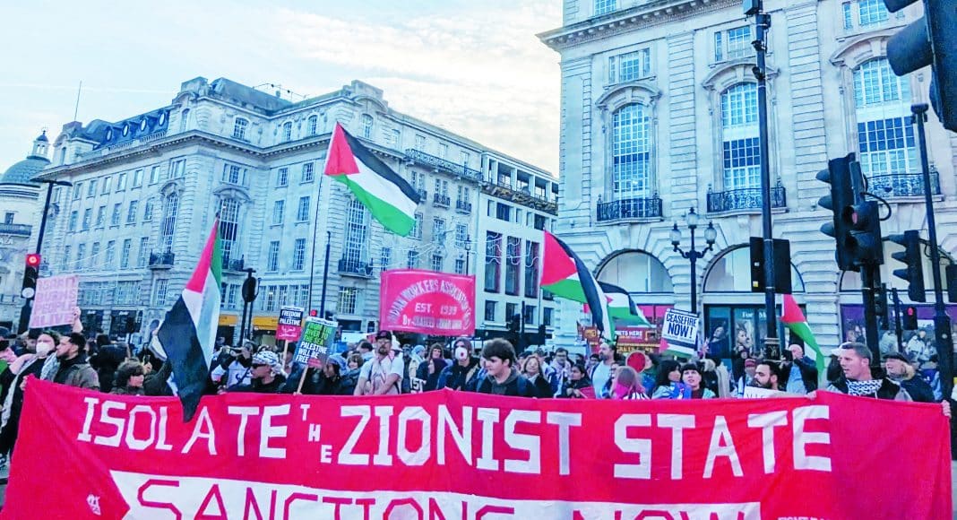 London RCG/FRFI contingent on the national march for Palestine, 30 November 2024 (Photo: FRFI). Protesters holding a large red banner reading 'Isolate the Zionist state, sanctions now' march along a London street with Palestine flags.