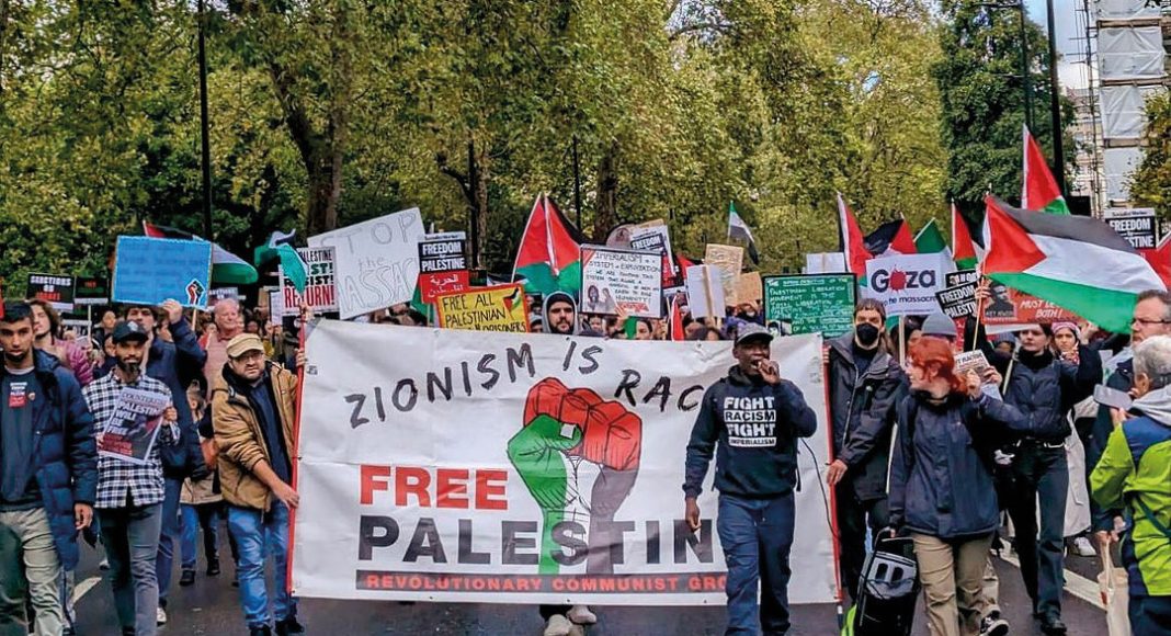 RCG protest in support of Palestine. A man speaks on a microphone in front of a benner reading 'Zionism is racism, free Palestine' marching in a crowd. (Photo: FRFI)