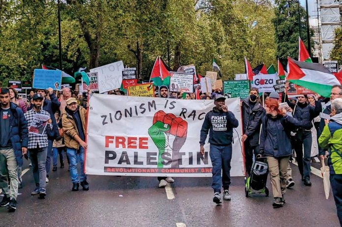 RCG protest in support of Palestine. A man speaks on a microphone in front of a benner reading 'Zionism is racism, free Palestine' marching in a crowd. (Photo: FRFI)