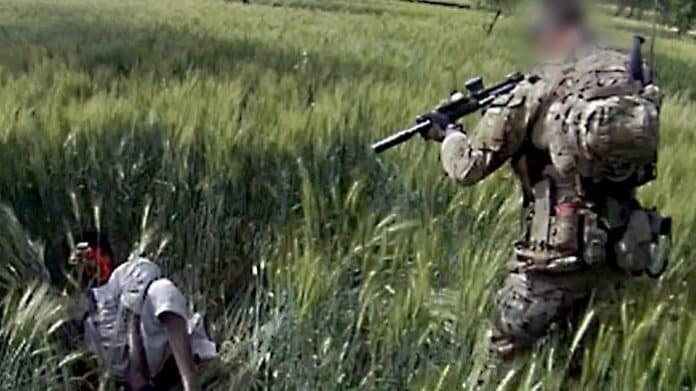 A solider aims a gun at an injured person lying in a field.