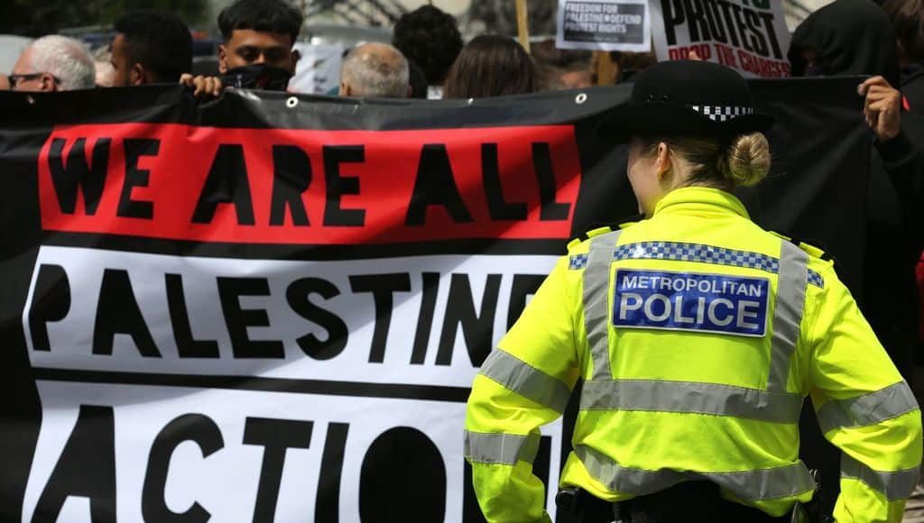 A police officer stands in front of protesters holding a red, white and black banner reading 'we are all Palestine Action'