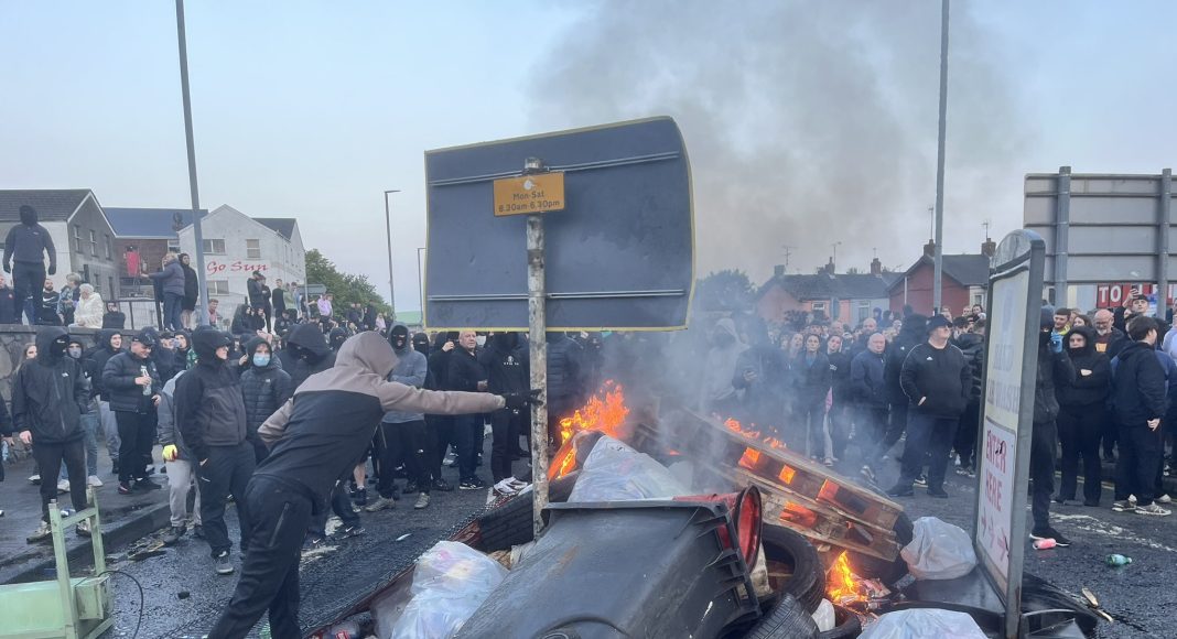 A man throws refuse onto a bonfire in a street in the north of Ireland while a crowd looks on (photo: The Flare/X)