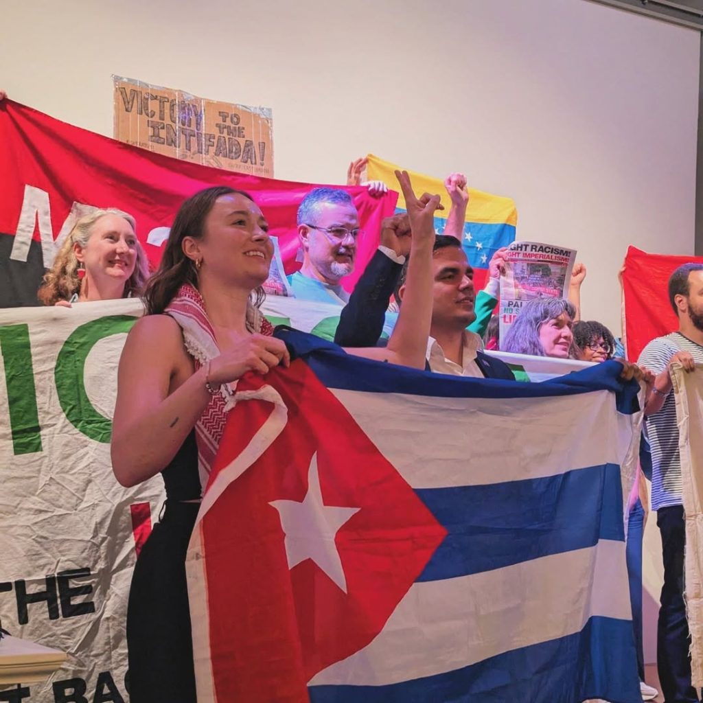 Solidarity with Cuba meeting at Bolivar Hall, 16 July 2025. Participants stand together on the stage holding up banners and flags with raised fists