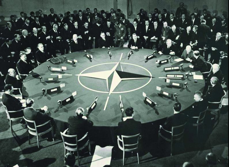 Men in suits sit around a round table with the NATO symbol in its centre, black and white photo.