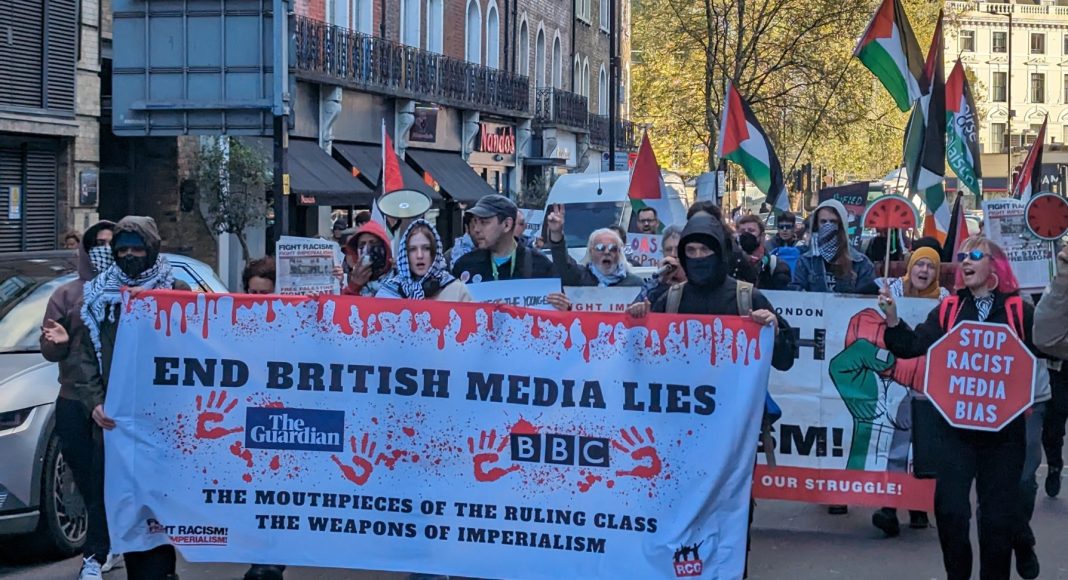 Protesters march on a London street carrying a banner reading 'End British media lies - the mouthpieces of the ruling class, the weapons of imperialism' with Guardian and BBC logos on it. Palestine flags are in the background