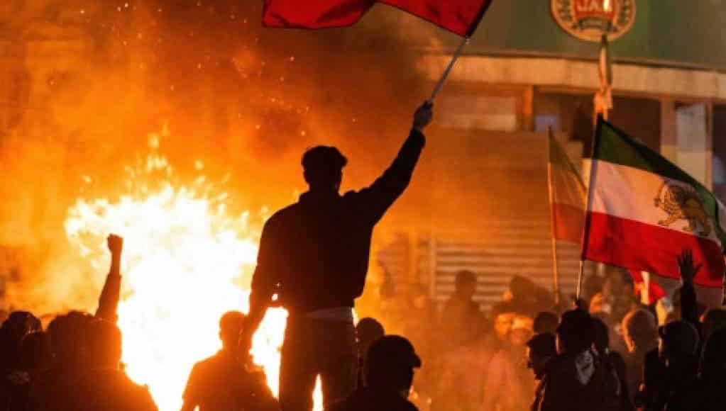 Demonstrator silhouetted against a street fire waving the Lion and Sun flag, 9 January 2026 (Photo: @gghamari/X)