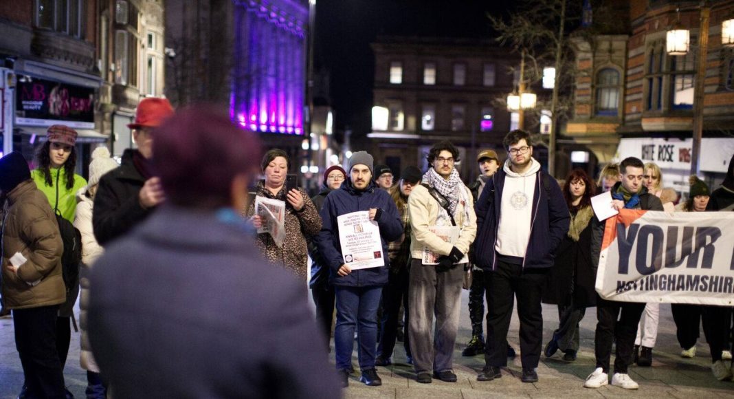 Vigil organised by Nottingham Stop Trump. People stand holding placards listening to a speaker in the foreground, in Nottingham at night. (photo: Cris Tregenza)