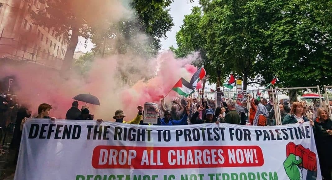 Protesters march on a London street carrying a banner reading 'defend the right to protest for Palestine/drop all charges now/resistance is not terrorism/Fight Racism! Fight Imperialism!'.
