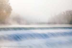 Ein foto vom Lechwehr in Landsberg am Lech im Morgennebel mit der Karolinenbrücke im Hintergrund