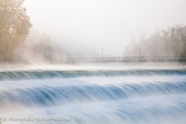 Ein foto vom Lechwehr in Landsberg am Lech im Morgennebel mit der Karolinenbrücke im Hintergrund