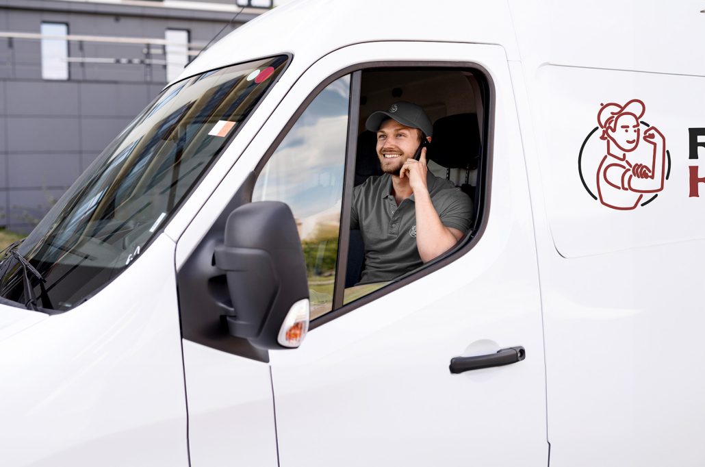 Lächelnder Umzugshelfer der Firma Rümpelkraft telefoniert im weißen Transporter mit Firmenlogo auf der Tür, bereit für den nächsten Einsatz.