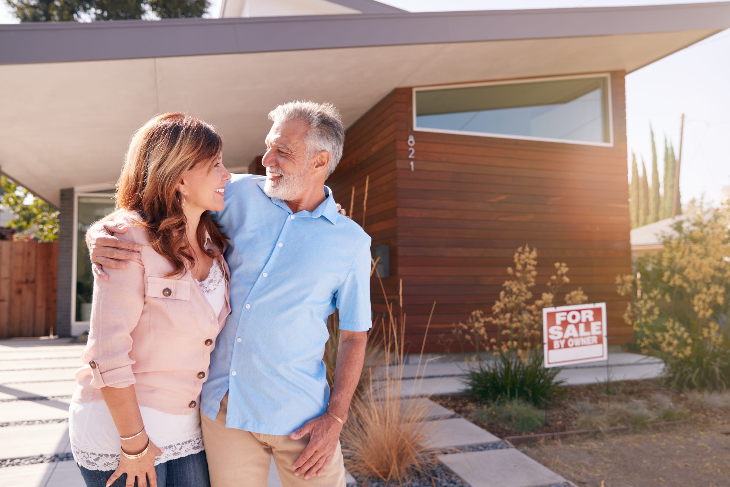 Senior Couple Standing Outdoors In Front Of House With For Sale Sign In Garden