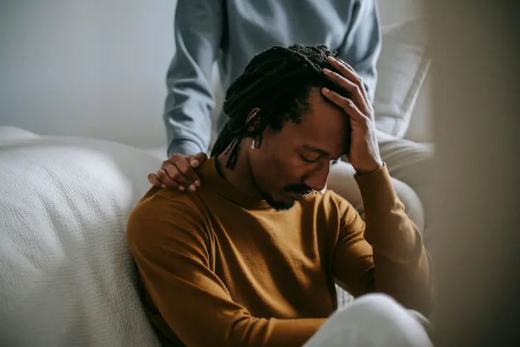 A man, holding up a hand to his head, being comforted by another person.