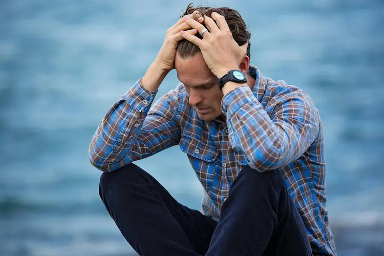 A man, sitting by a body of water. holding his head in his hands.
