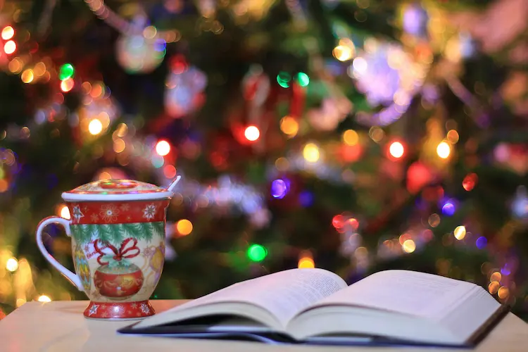 A festive mug and an open book on a table in front of a lit Christmas tree