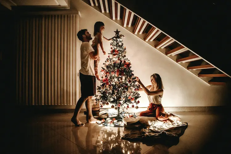 A man lifting a small child to decorate a Christmas tree while a woman arranges presents underneath