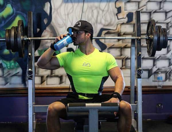 Sports coach Julien Quaglierini consumes a shaker while sitting on a weight bench in a fitness center.