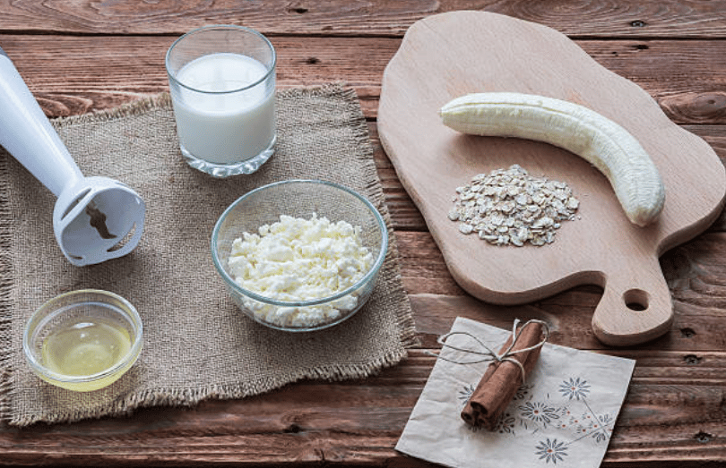 A table with different ingredients to prepare a home-made gainer: oatmeal, milk, whey protein and banana.