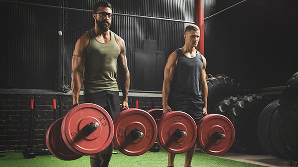 2 hommes vêtus d'un débardeur réalisent l'exercice de la marche du fermier dans une salle de sport