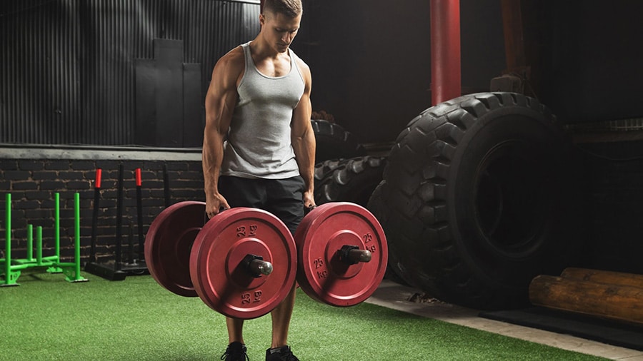 A man in a tank top performs the farmer walk exercise in a weight room