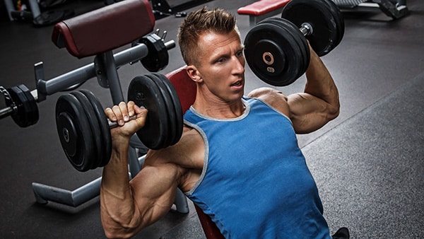 A man in a tank top performs a shoulder exercise at the gym.