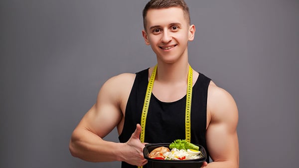 A man in a tank top, with a tape measure around his neck, gives a thumbs up as he presents a balanced meal.