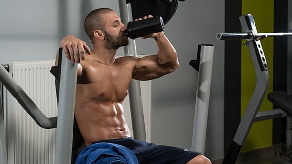 A shirtless man drinks from a shaker during a workout at the gym.