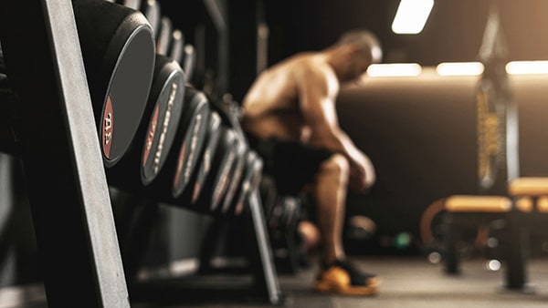 A weightlifter sitting on a bench in a gym, with dumbbells in the foreground.