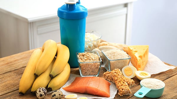 A table with many foods: bananas, salmon, whey protein, cheese and a shaker for supplements.