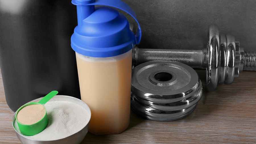 A shaker composed of a mass gainer, a bowl with protein powder and a dumbbell.