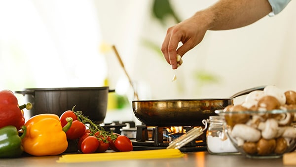 A man in the kitchen adds ingredients to a pan, with vegetables placed in front of him.