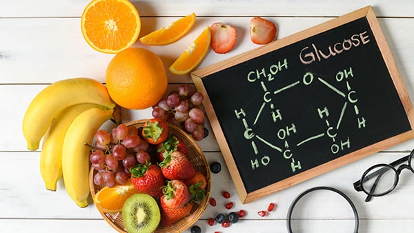 A table with fruits and vegetables (banana, orange, strawberry, kiwi) and a blackboard representing glucose.