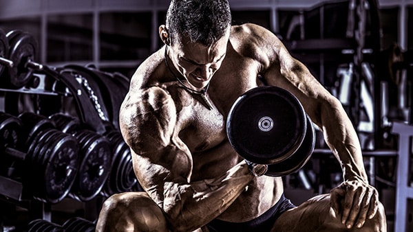 A muscular, shirtless man performs a bodybuilding exercise to build biceps with the rest pause technique.